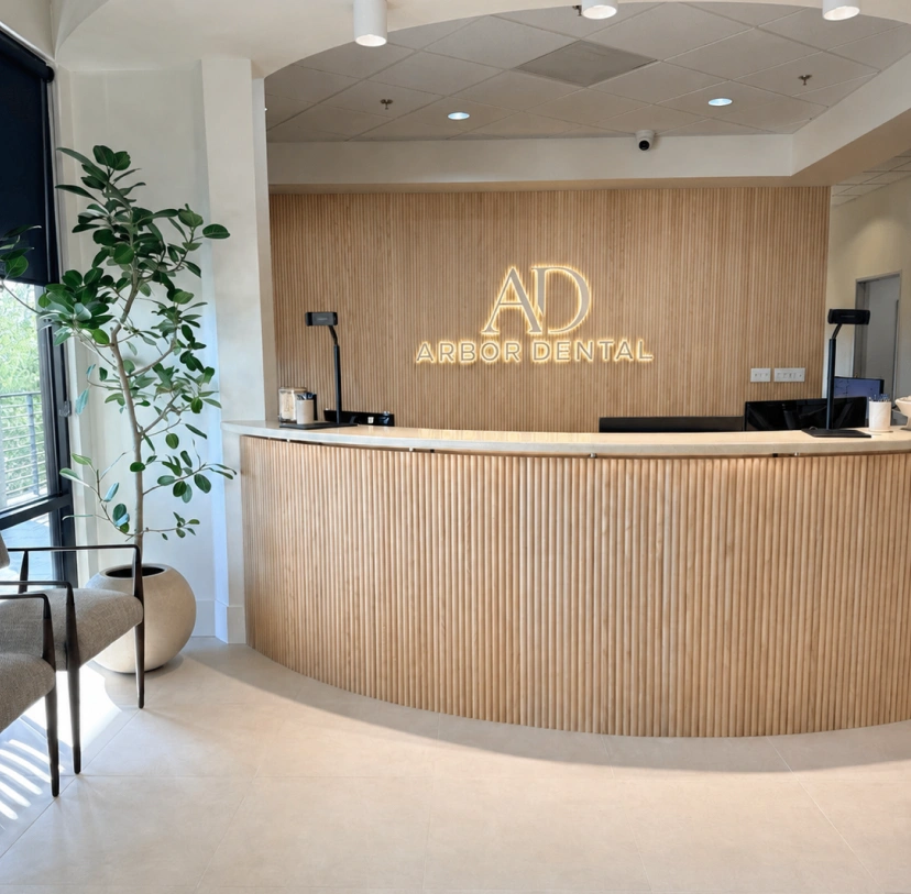 Brightly lit reception area at Arbor Dental featuring a wide, curved reception desk made of light-wood vertical slats with a white countertop. The matching back wall displays a glowing, backlit sign with the letters "AD" above the words "ARBOR DENTAL". To the left, a tall potted ficus tree stands near a large window and modern upholstered waiting chairs, under a tiled ceiling with recessed lighting.